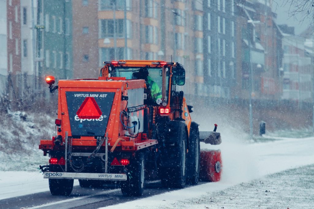 Orange snowplow in action clearing a snow-covered street during a winter snowfall.